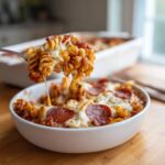 A fork lifting a cheesy, saucy portion of pizza pasta bake out of a white bowl, showing melted cheese strings.