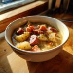 A close-up of a creamy potato and sausage chowder in a white bowl, garnished with herbs.
