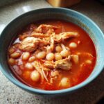 Close-up of a vibrant red bowl of chicken pozole featuring shredded chicken and large white hominy kernels.