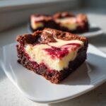 A close-up of a decadent red velvet cheesecake brownies square with swirled cream cheese topping on a white plate.