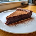 A decadent slice of flourless chocolate torte dusted heavily with cocoa powder, served on a white plate.
