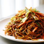 Close-up of a mound of glossy, dark brown fried noodles mixed with julienned carrots and cabbage.