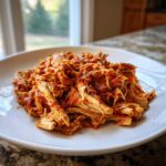 A close-up of shredded chicken tinga coated in rich, red sauce served on a white plate.