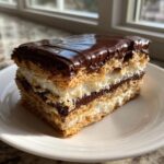 A cross-section slice of chocolate eclair cake showing layers of graham cracker crust, vanilla cream, and chocolate filling, topped with shiny chocolate glaze.