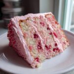 A moist slice of 1-layer cherry cake featuring visible red cherry pieces throughout the light sponge and covered in pink frosting.