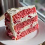 A tall slice of moist, pink strawberry cake layered with white frosting and visible pieces of strawberry.