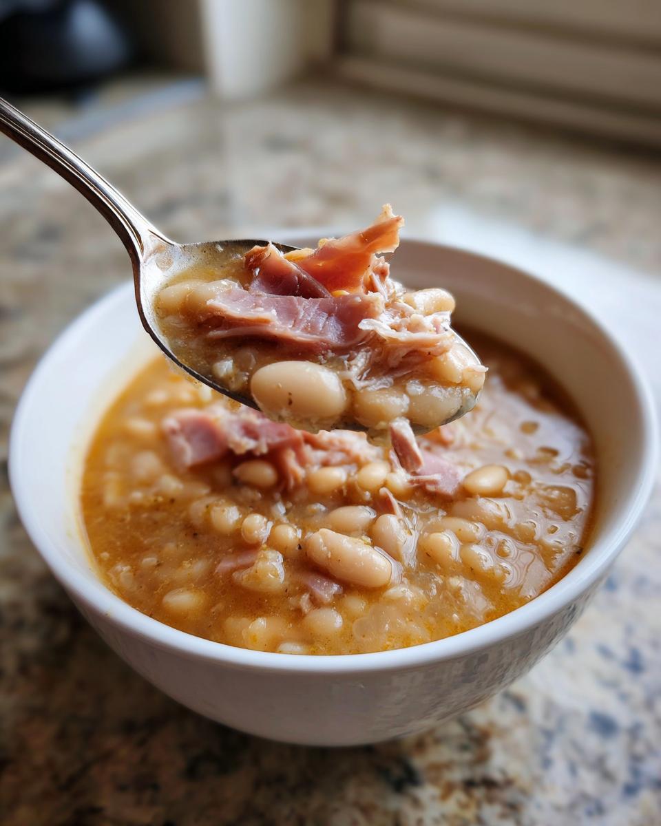A spoonful of thick navy bean soup loaded with white beans and shredded ham being lifted from a white bowl.