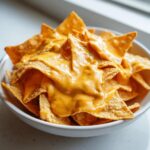 Close-up of tortilla chips generously covered in smooth, bright orange nacho cheese dip in a white bowl.
