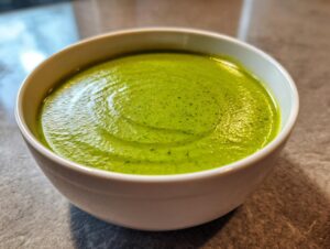A close-up of a bowl filled with creamy, vibrant green zucchini soup, showing swirls on the surface.